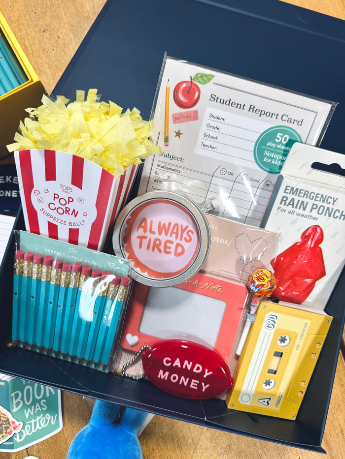 A curated student gift box or surprise mailer filled with themed items including a red-and-white striped popcorn bag, teal pencils, a pink card holder, a red 'Candy Money' charm, a yellow cassette tape, a red rain poncho, and a student report card, all arranged inside a dark box.