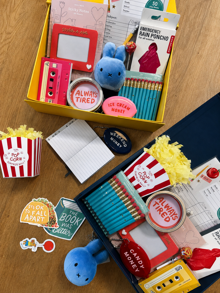 A curated back to school themed stationery and surprises gift set for kids, featuring two open boxes with notebooks, pencils, stickers, a blue plush squish toy, a retro mixtape notebok, a rain poncho, and popcorn-themed surprise ball.
