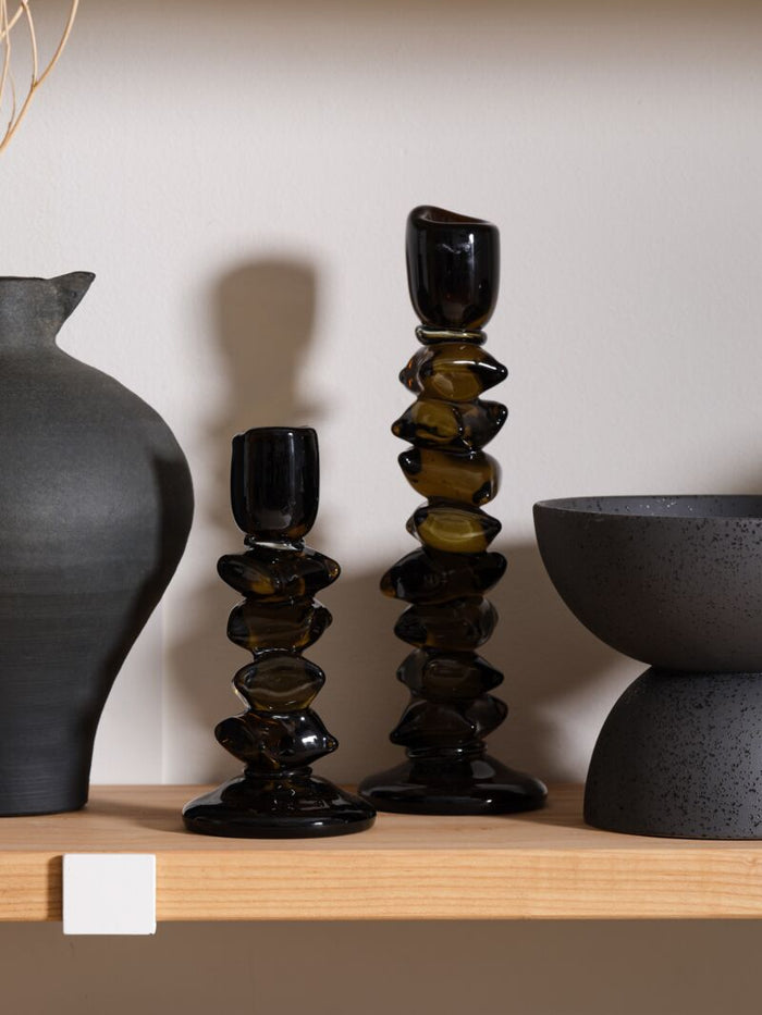 Two dark green sculptural glass candleholders displayed on a wooden shelf with ceramic decor pieces.