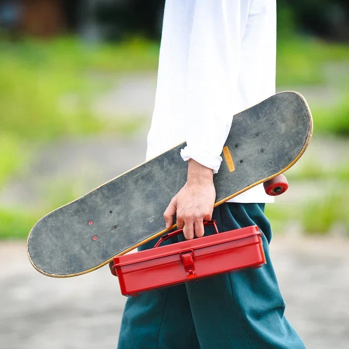 Person carrying a red metal toolbox and a skateboard outdoors, shown from the waist down.