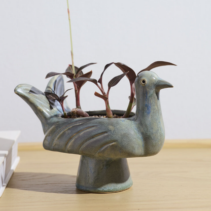 A ceramic bird-shaped planter in a muted blue-green glaze, holding several small dark-leafed plants, set on a light wood surface against a plain light gray wall.